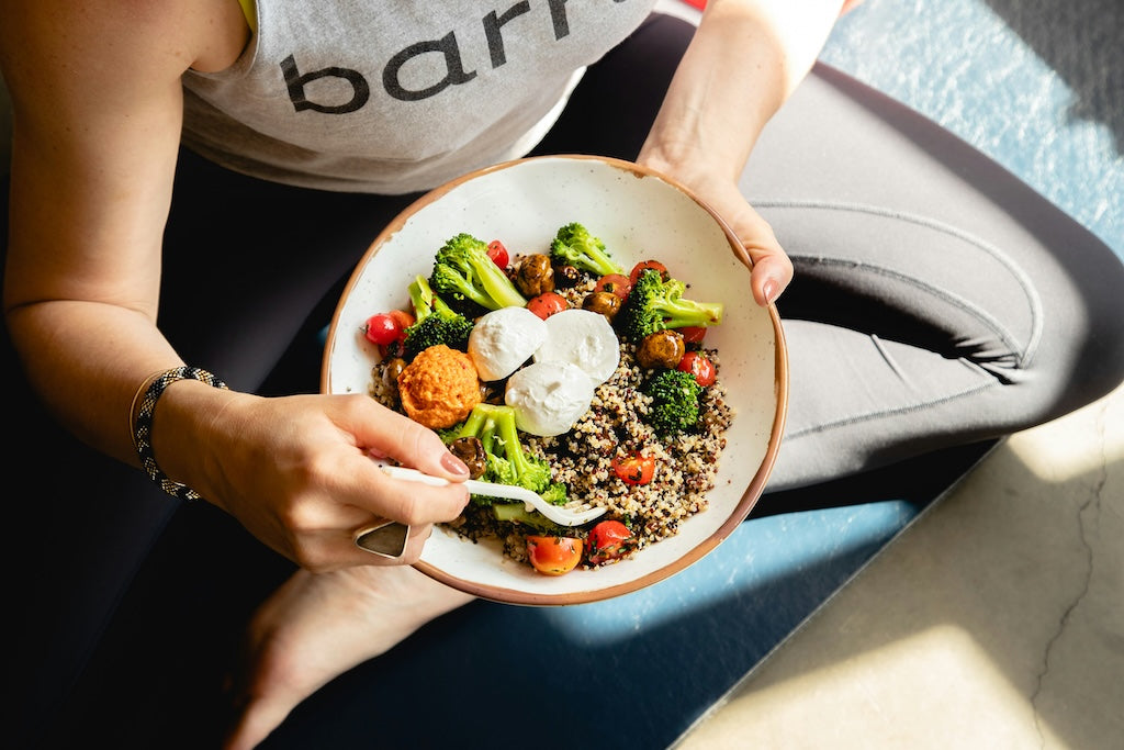 Une personne en tenue de sport est assise en tailleur, tenant un bol de quinoa, de brocoli, de tomates cerises et d'œufs pochés, et mange avec une fourchette.
