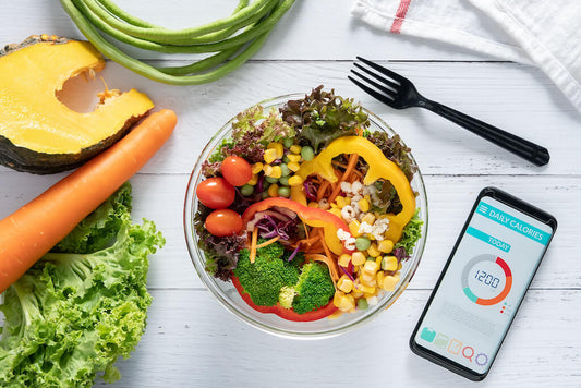 Una ciotola di insalata mista e verdure accanto a uno smartphone con un contatore giornaliero di calorie, posta su un tavolo di legno bianco con frutta e verdura fresca intorno.