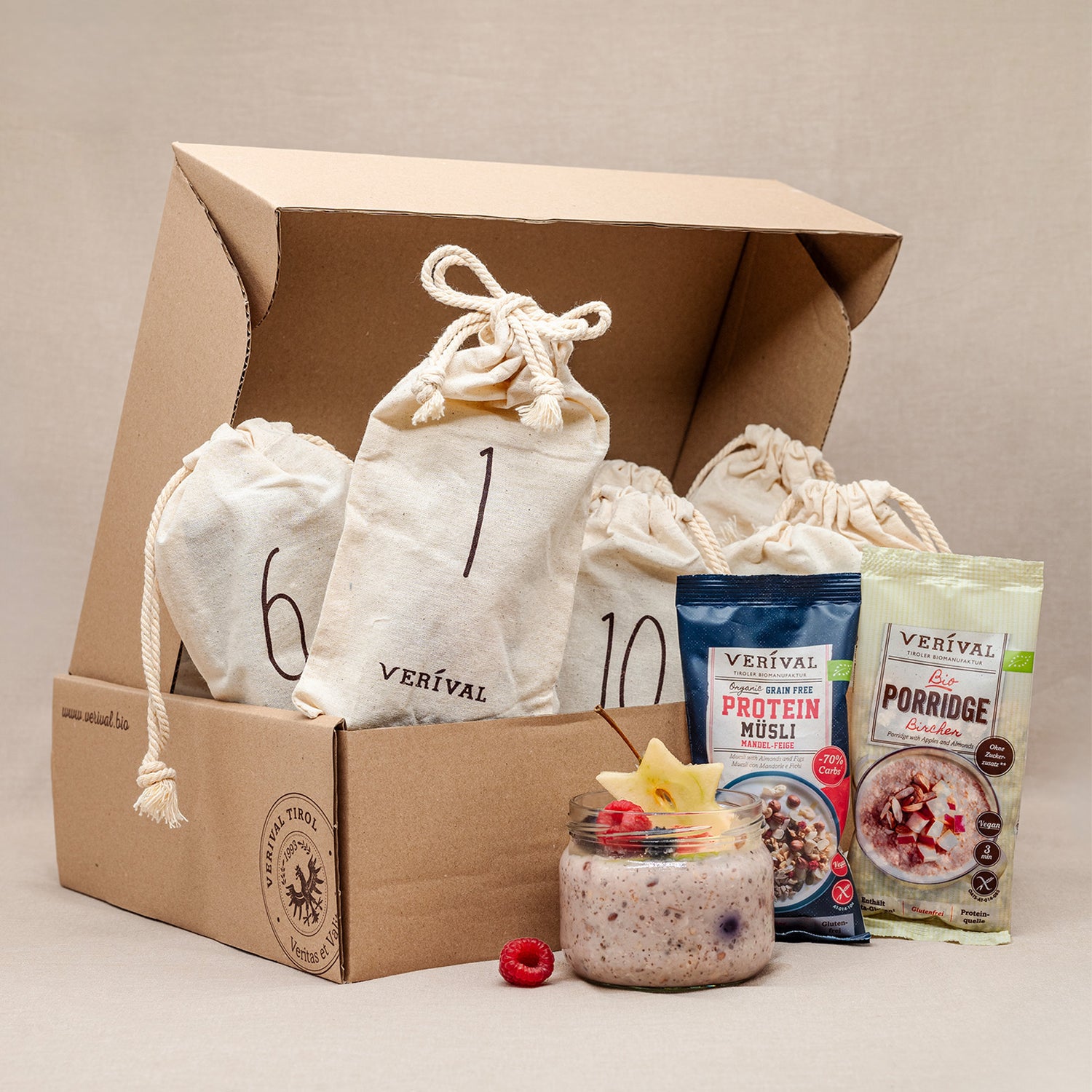 Eine Pappschachtel mit nummerierten Stoffbeuteln, zwei Päckchen mit der Aufschrift Protein Müsli und Porridge und ein Glas Müsli mit Früchten, alle mit der Marke Verival.