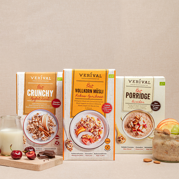 Three cereal boxes labeled Crunchy, Wholegrain Muesli, and Porridge are presented with a glass, cherries, and almonds on a beige background.