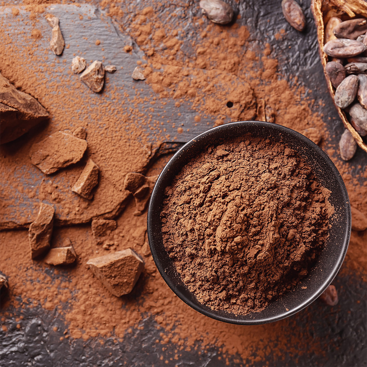 A black bowl filled with cocoa powder stands on a dark surface, surrounded by scattered cocoa powder, cocoa beans and chocolate pieces.