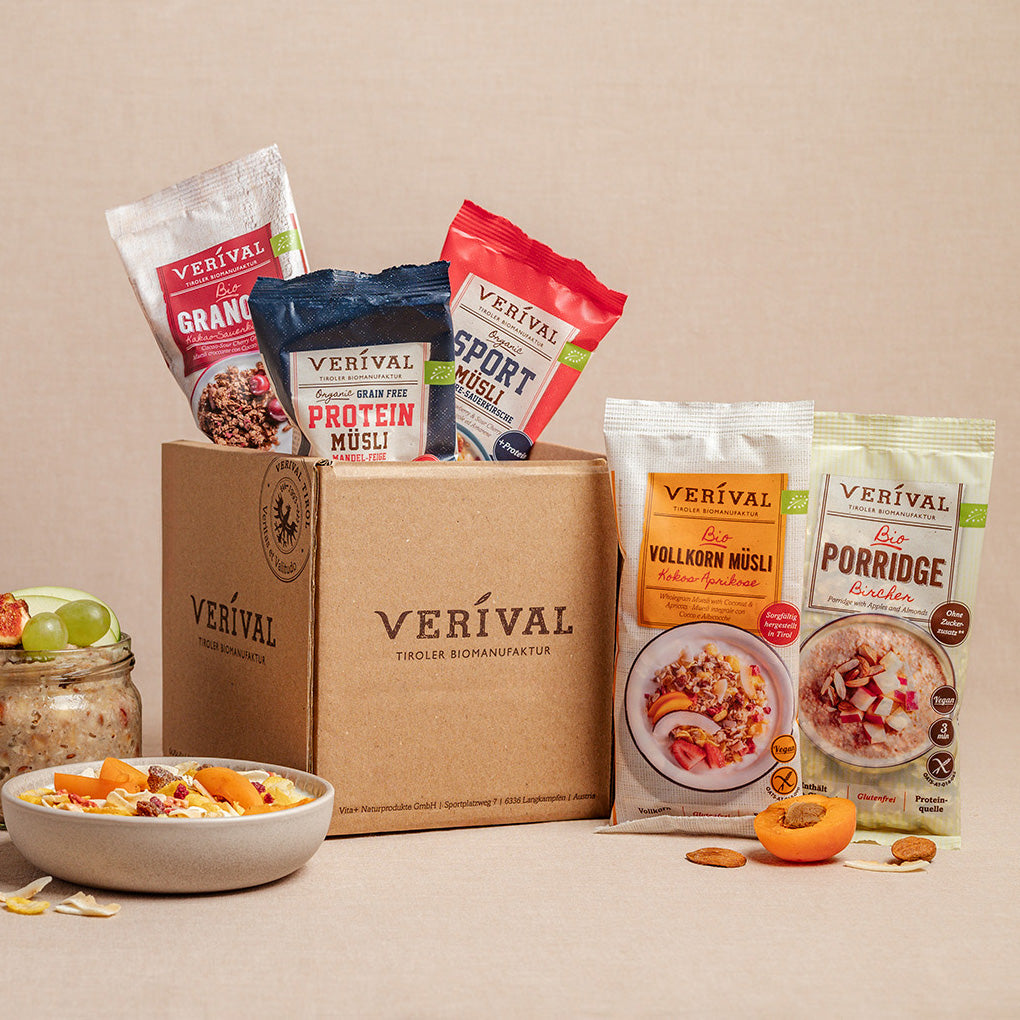 A box with the Verival logo, surrounded by five different muesli and porridge packets, in front of which are two bowls of prepared muesli and fruit.