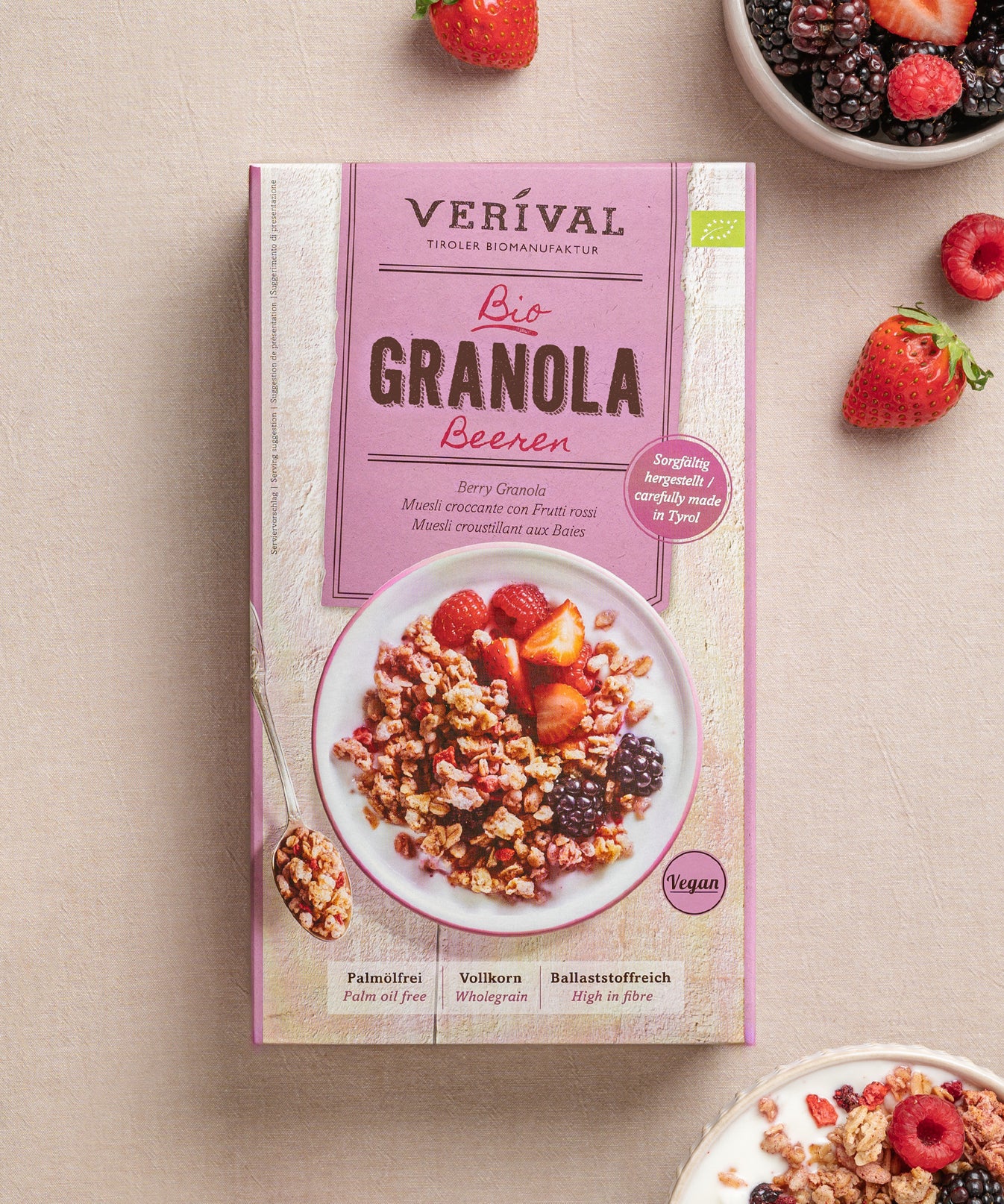 Eine Packung Beeren-Granola von Verival Bio steht neben einer Schüssel mit knusprigem Granola, Erdbeeren und Brombeeren. Reich an Ballaststoffen bietet dieser vegane Leckerbissen ein nahrhaftes Frühstück oder eine Snack-Option.