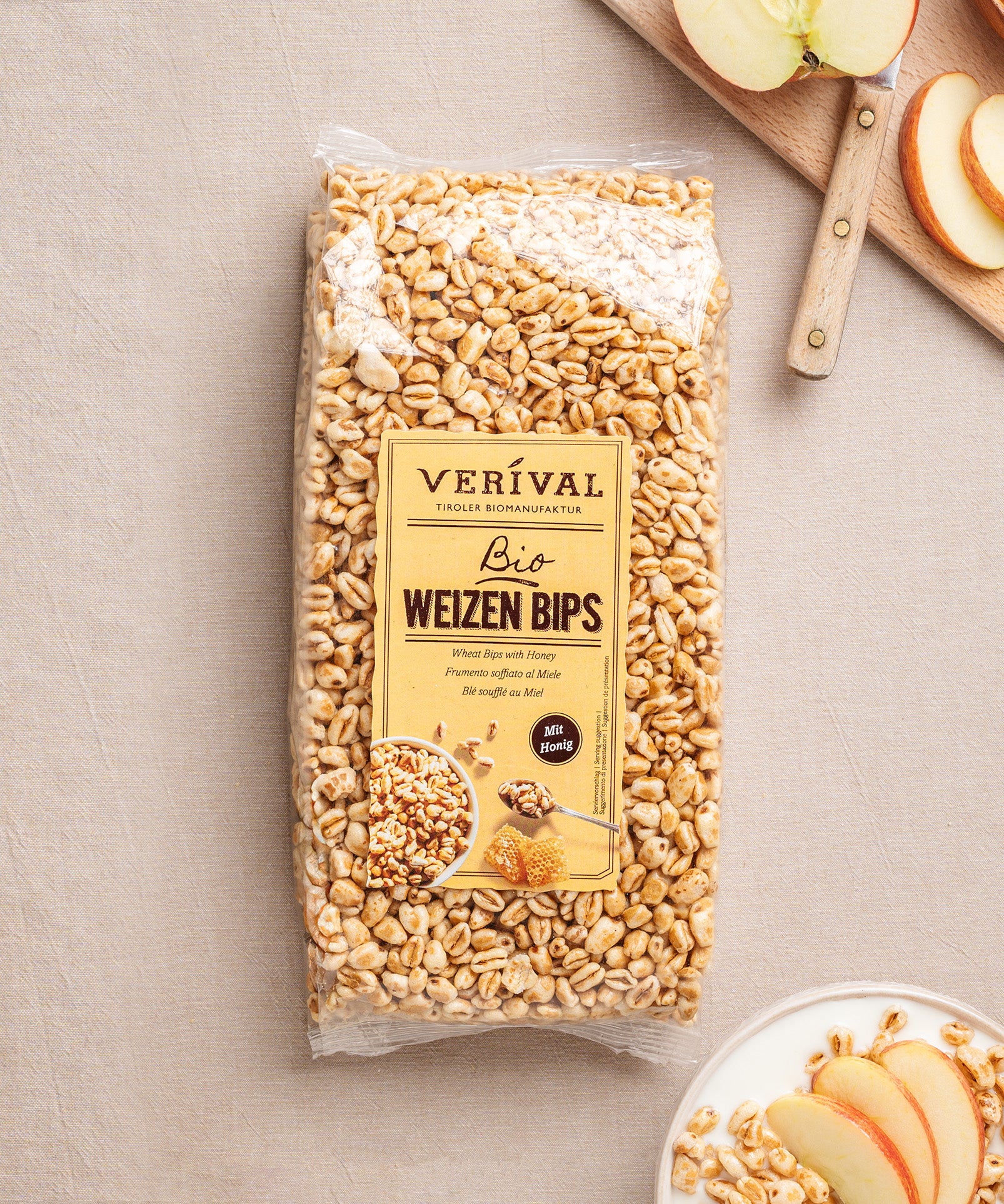 Eine Packung Weizen Bips mit Honig von Verival Bio wird elegant auf einer beigen Oberfläche mit Apfelscheiben auf einem Teller und einem Schneidebrett präsentiert.