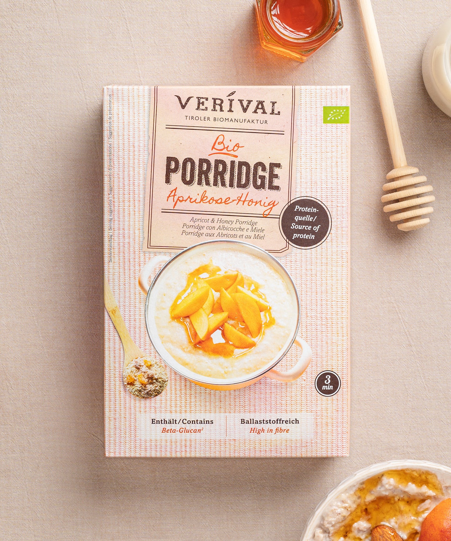 Der Aprikosen-Honig Porridge von Verival Bio mit hohem Protein- und Ballaststoffgehalt wird kunstvoll mit Aprikosenscheiben präsentiert. Ein Honigglas und ein Schöpflöffel runden den laktosefreien Genuss ab.