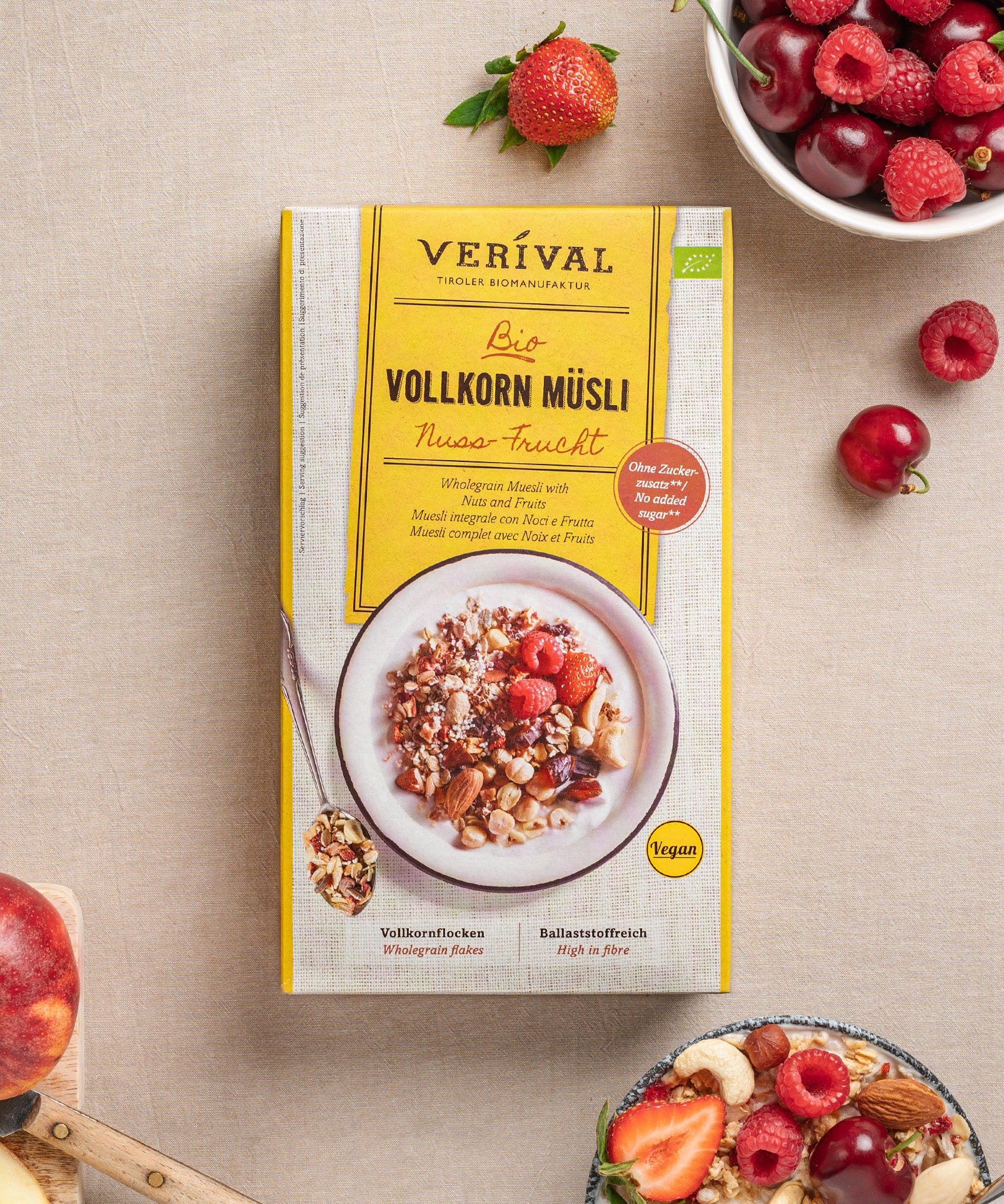 Eine Packung Nuss-Frucht Vollkorn Müsli von Verival Bio steht auf einem Tisch mit verschiedenen Früchten, geschnittenen Pfirsichen und Kirschen. Diese vegane Frühstücksoption ist köstlich und ballaststoffreich – perfekt für alle, die Wert auf eine nahrhafte Alternative legen.
