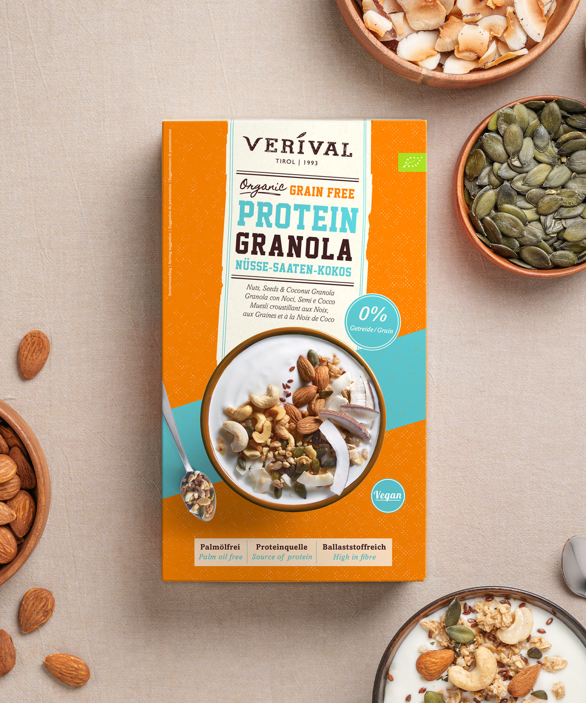 Eine Packung Bio Nuss-Kokos Grain Free Protein Granola von Verival Bio GmbH, umgeben von kleinen Schälchen mit Mandeln, Kürbiskernen und Kokosflocken sowie einem Schälchen Joghurt, das mit dem pflanzlichen Protein-Müsli garniert ist.