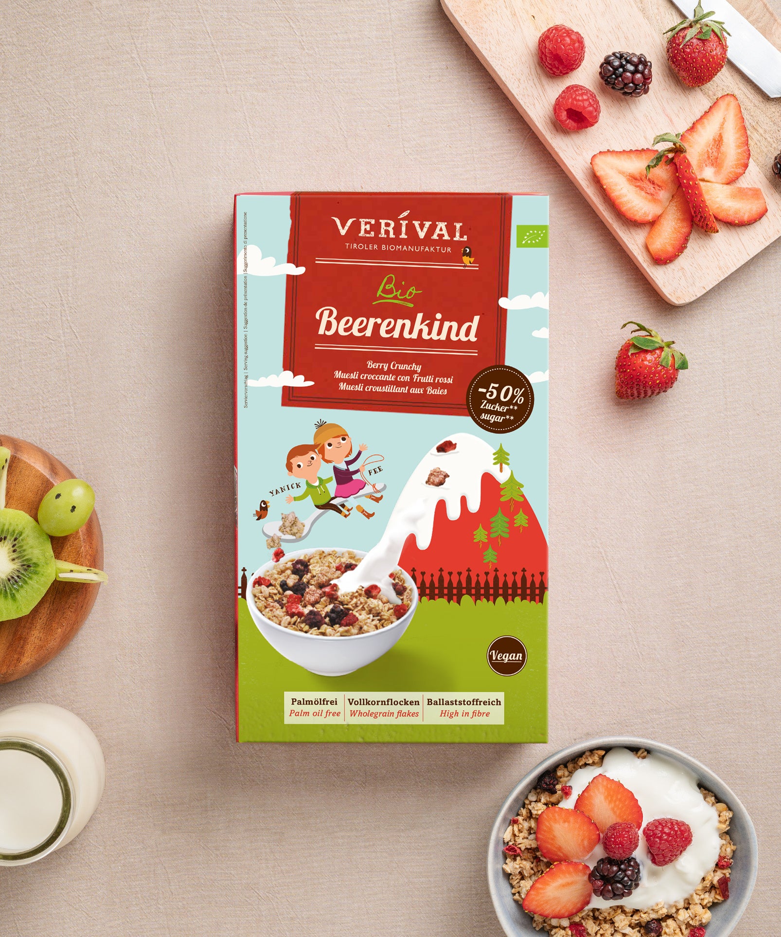 Eine Packung veganes Beerenkind-Müsli von Verival Bio steht auf dem Tisch, neben einer Schüssel mit der knusprigen Mischung, frischen Beeren, Kiwischeiben und einem Glas Milch. Vollgepackt mit Ballaststoffen ist es ein köstlich nahrhafter Start in den Tag.
