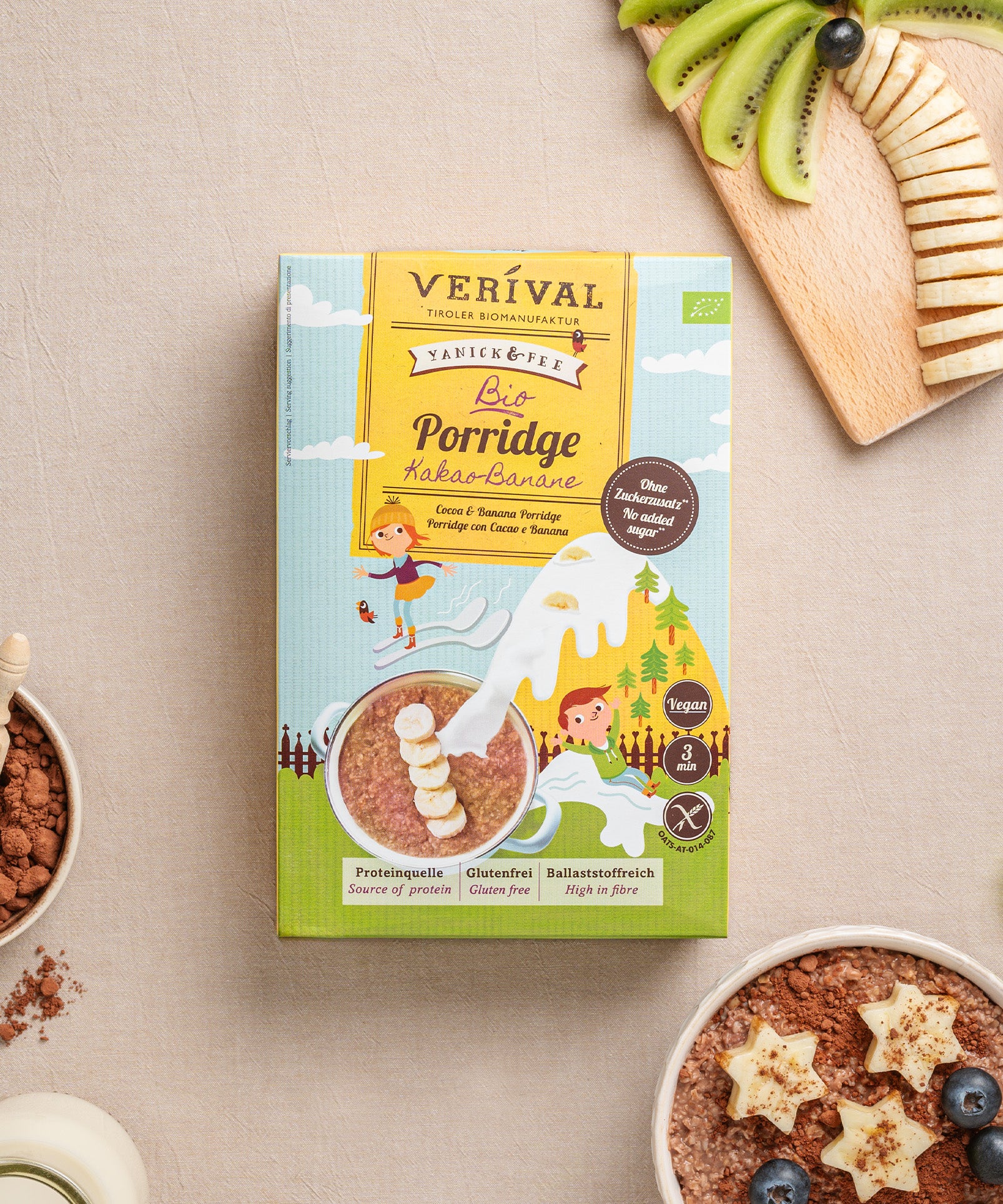 Eine Packung Verival Bio Kinderporridge Kakao-Banane steht neben einer Schüssel cremigem Porridge mit Kiwi- und Bananenscheiben sowie einer Kokosnussschale mit Nüssen auf dem Tisch. Dieser glutenfreie Genuss steckt voller Ballaststoffe für einen nahrhaften Start in den Tag.