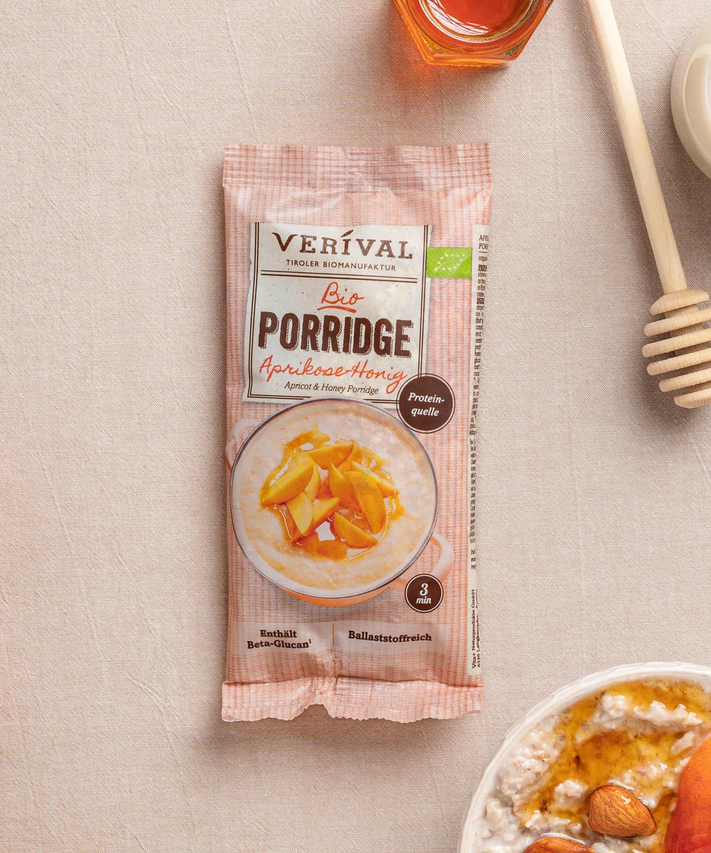 Eine Einzeportinspackung Aprikosen-Honig-Porridge von Verival Bio  liegt auf einer beigen Oberfläche neben einer Schüssel cremigen Porridges mit orangefarbenen Fruchtstücken und einem Honiglöffel.