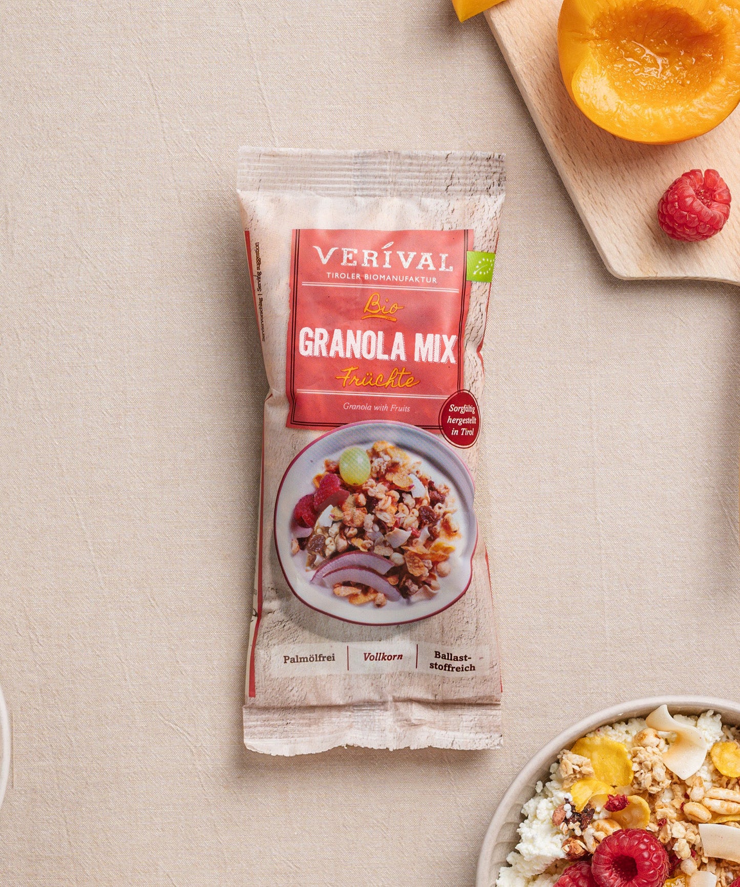 Eine Packung des ballaststoffreichen Granola Mix Früchte von Verival Bio GmbH liegt auf einem beigen Untergrund. Daneben stehen eine Müslischale mit Früchten sowie ein Holzbrett mit Himbeeren und einer halben Aprikose.