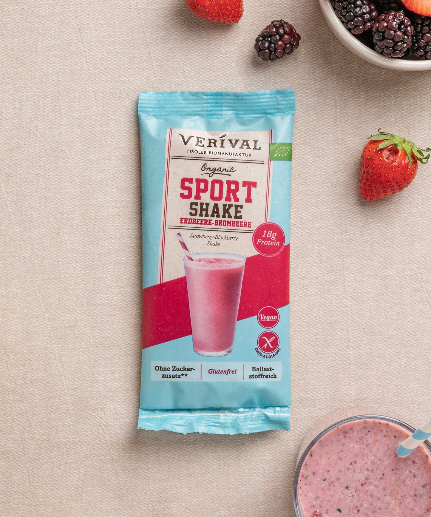 Eine Einzelportionspackung vom Sport Shake Erdbeere-Brombeere von Verival Bio ist umgeben von Erdbeeren, Brombeeren und dem Shake in einem Glas auf einem beigen Untergrund.