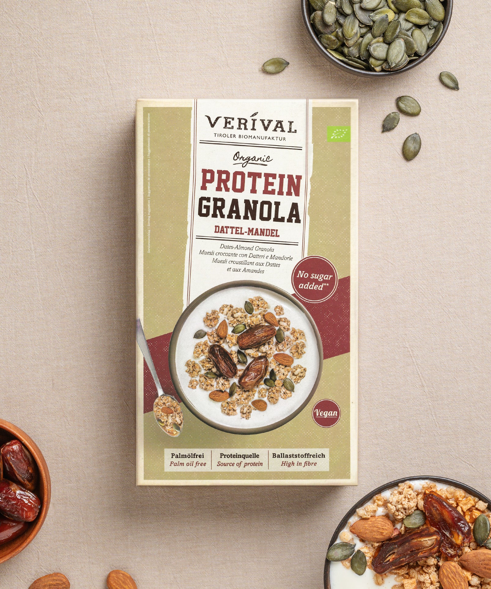 Eine Packung Protein-Granola Dattel-Mandel von Verival Bio, reich an pflanzlichen Proteinen, steht auf einer Tischdecke, umgeben von Schüsseln mit dem ballaststoffreichen Granola, umgeben von verstreuten Mandeln und Datteln.