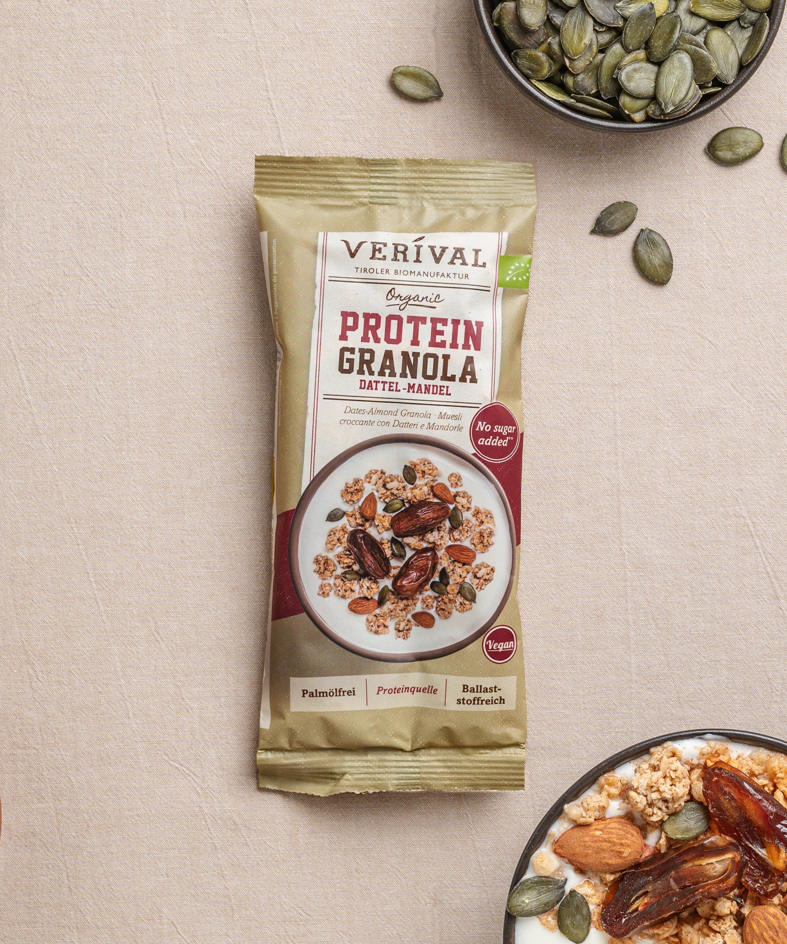 Eine Enzelportionspackung Protein-Granola Dattel-Mandel von Verival Bio steht neben einer Joghurtschale mit Granola, Mandeln, Datteln und Kürbiskernen in einer kleinen Schale.