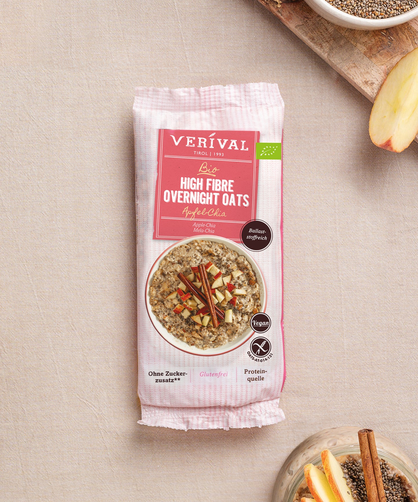 Eine Packung Bio High Fibre Overnight Oats Apfel-Chia der Verival Bio GmbH steht auf einer beigen Fläche, daneben befinden sich Schalen mit Chiasamen und Apfelscheiben, die auf dieses ballaststoffreiche Frühstück ohne Zuckerzusatz hinweisen.