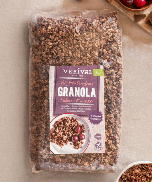 Eine Packung Verival Bio GmbH Bio Kakao-Kirsche Granola glutenfrei steht auf einer beigen Fläche neben einer Schale und Kirschen. Dieses ballaststoffreiche Granola ist perfekt für ein gesundes Frühstück.