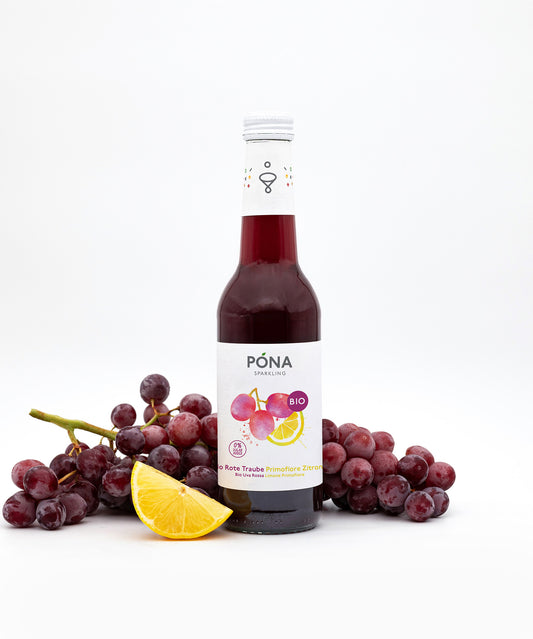 Eine Flasche PONA Rote Traube-Primofiore Zitrone der Wonderful Biodrinks GmbH steht neben roten Trauben und einer Zitrone. Das Etikett mit Trauben- und Zitronenmotiven präsentiert den Bio-Fruchtsaft mit Roter Traube, Zitrone und Eisenkraut auf weißem Hintergrund.