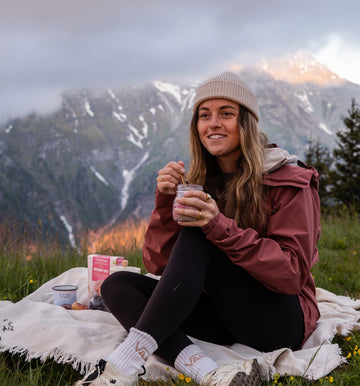 Eine Person sitzt auf einer Decke in den Tiroler Bergen, hält ein Glas mit Porridge in der Hand und lächelt. Auf der Decke liegen eine Schachtel Overnight Oats von Verival Bio und etwas Obst. Im Hintergrund erheben sich majestätisch wolkenverhangene Gipfel der tiroler Berge.