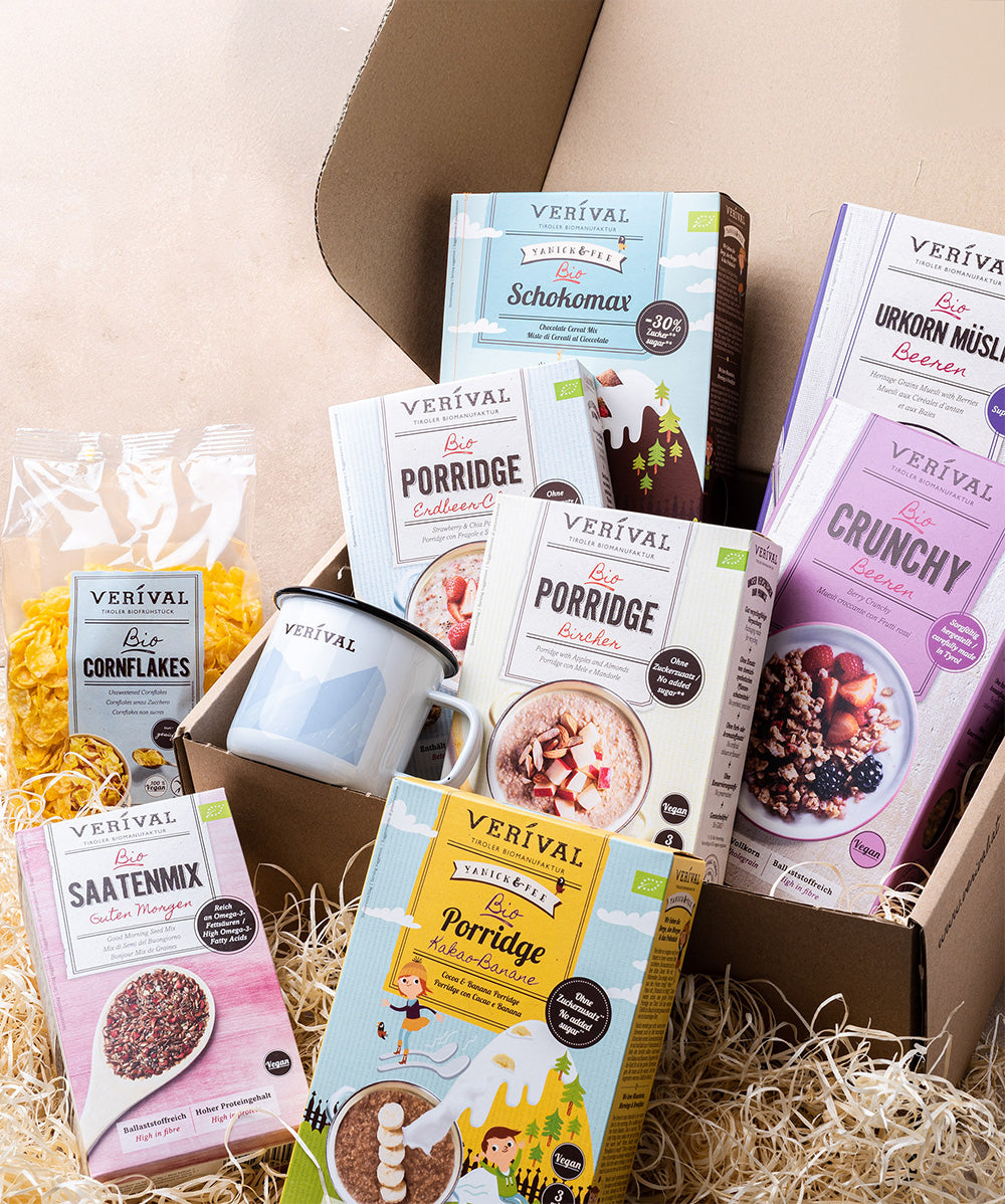 Die Geschenkverpackung für Set "Best Breakfast Friends" von Verival Bio GmbH zeigt Porridge, Müsli, Cornflakes, eine Saatenmischung und einen Becher, allesamt ansprechend auf strohhalmartigem Verpackungsmaterial angeordnet.
