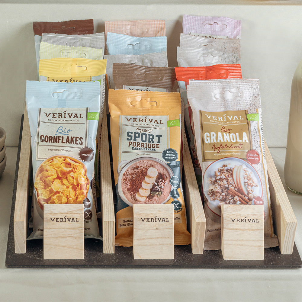 Eine Vielzahl von Verival Frühstücks-Cerealienpackungen, darunter Einzelportionen von Cornflakes, Porridge und Müsli, werden in Holzhaltern in einem Regal präsentiert.