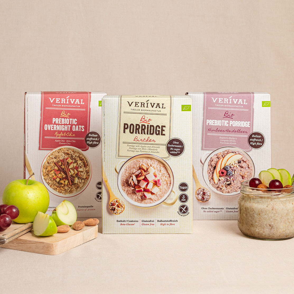 Drei Packungen Verival Frühstückscerealien – Prebiotic Overnight Oats, Bio Porridge und Prebiotic Porridge – präsentiert mit einer Schüssel Porridge, Apfelscheiben und Weintrauben davor.