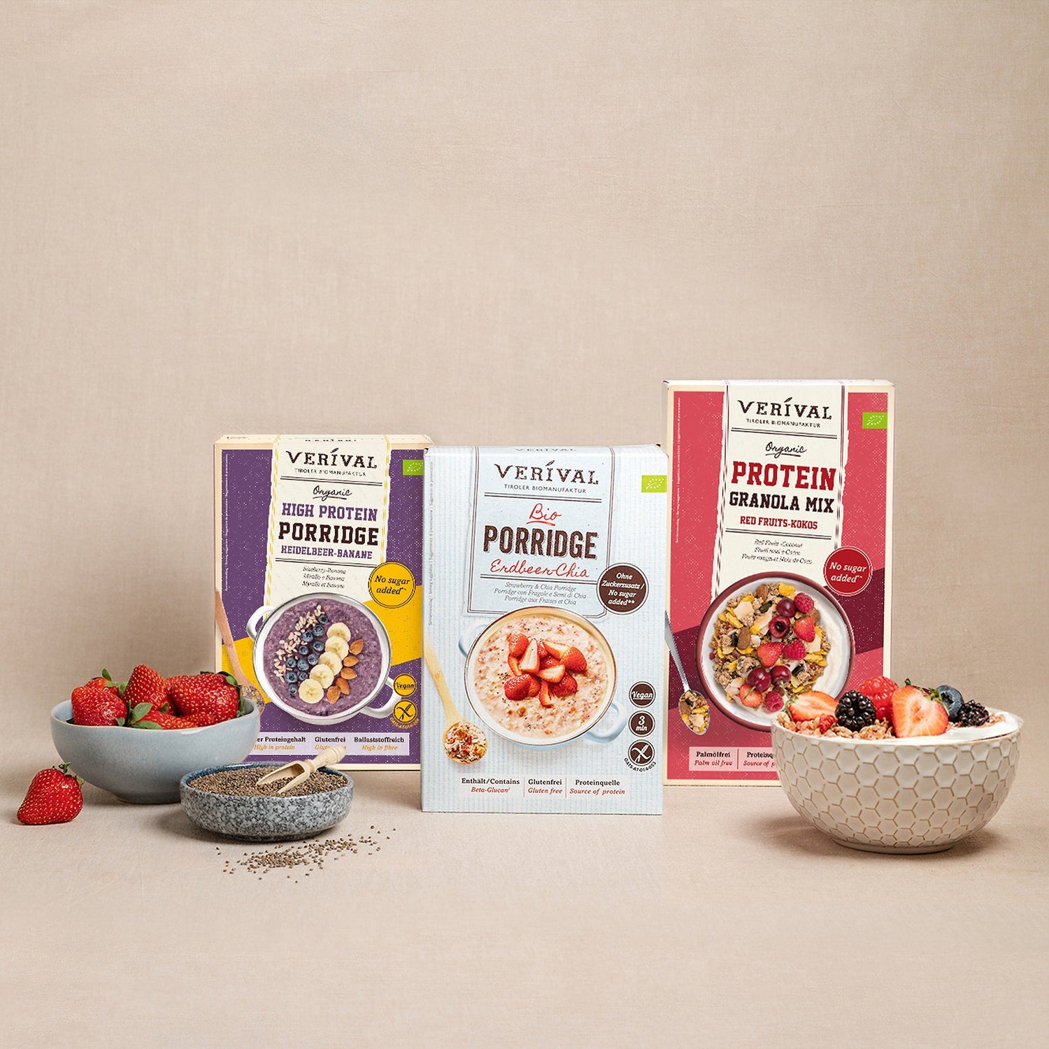 On display are three Verival porridge and granola boxes, surrounded by bowls of fresh berries and chia seeds on a neutral background.