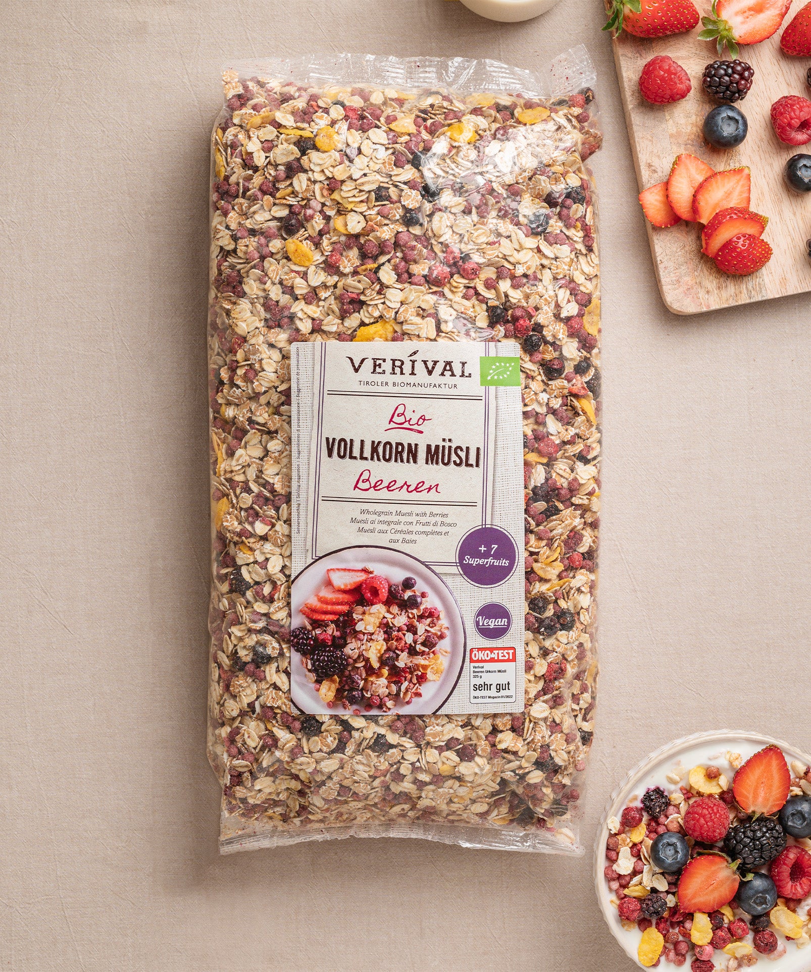 Auf beigen Untergrund steht eine Packung Beeren Vollkorn Müsli von Verival Bio. Daneben steht eine Schüssel mit dem ballaststoffreichen Müsli, garniert mit Beeren. Auf einem Holzbrett sind Früchte zu sehen: geschnittene Erdbeeren, Brombeeren und Himbeeren.