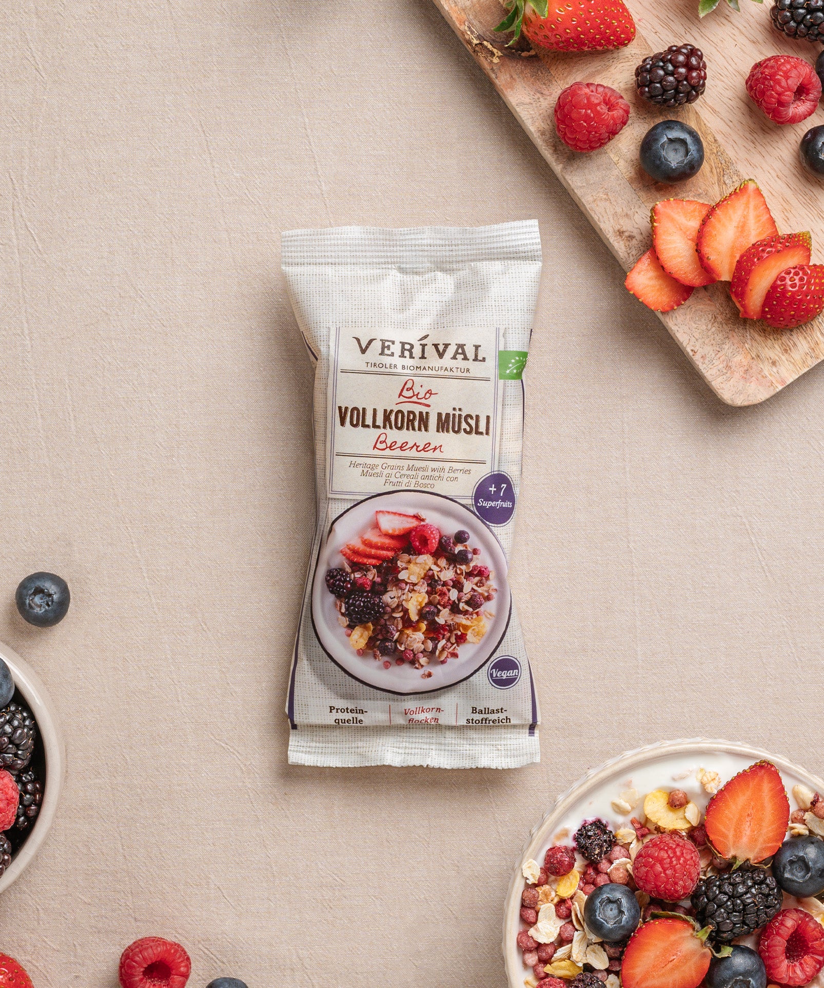 Ein Einzelportionspackung Beeren Vollkorn Müsli von Verival Bio, umgeben von Schalen mit diesem ballaststoffreichen Müsli und einem Holzbrett mit gemischten Beeren. 
