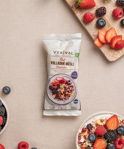 Ein Einzelportionspackung Beeren Vollkorn Müsli von Verival Bio, umgeben von Schalen mit diesem ballaststoffreichen Müsli und einem Holzbrett mit gemischten Beeren. 