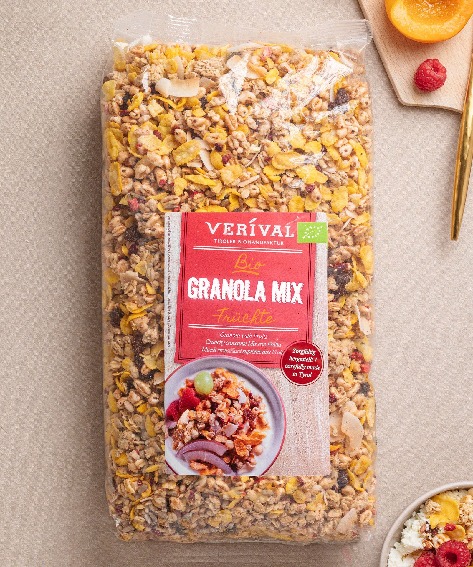 Eine Packung Granola Mix Früchte von Verival Bio mit dem roten Etikett, auf dem eine Schale ballaststoffreiches Granola abgebildet ist, umgeben von Orangen, Himbeeren und Heidelbeeren.