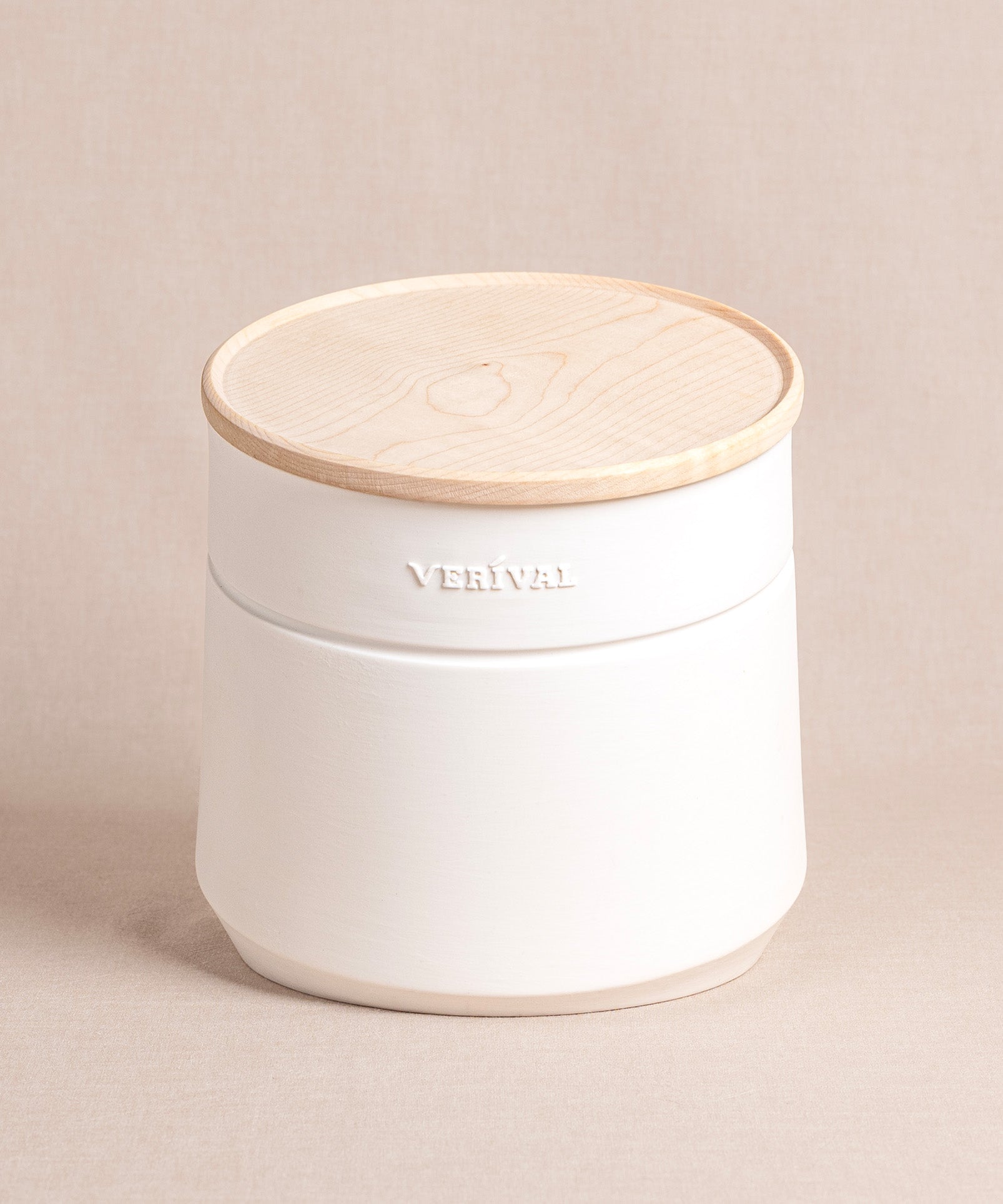 Der „Frühstücksbehälter für Porridge, Granola, Müsli & Co.“ von Verival Bio ist ein weißer Keramikbehälter mit einem Deckel aus Ahornholz auf beigem Untergrund, dessen elegantes Design durch die Prägung des Schriftzugs VERIVAL unterstrichen wird.