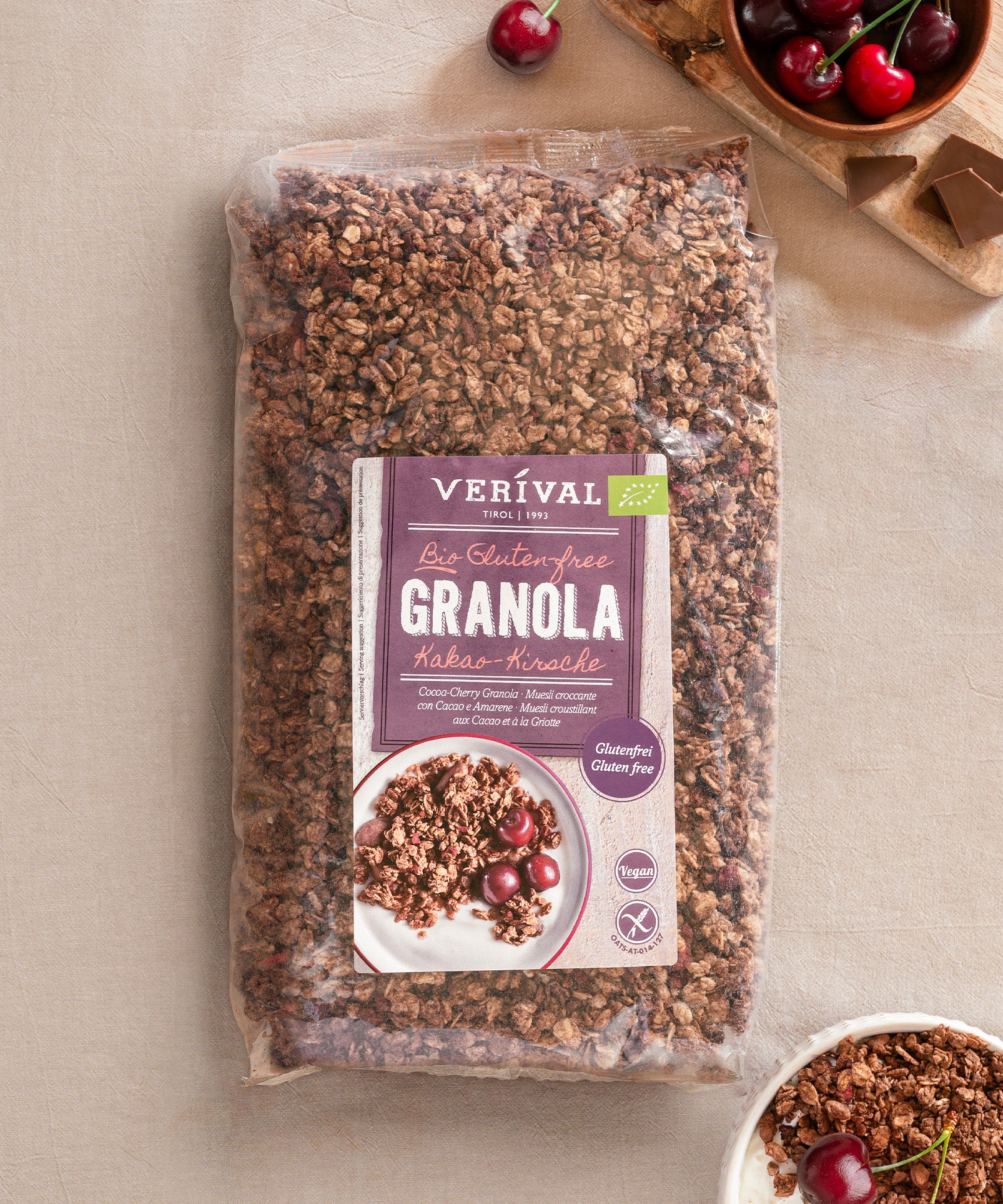 Eine Packung Verival Bio GmbH Bio Kakao-Kirsche Granola glutenfrei liegt auf einer beigen Fläche, daneben eine Schale mit dem Granola und frischen Kirschen.