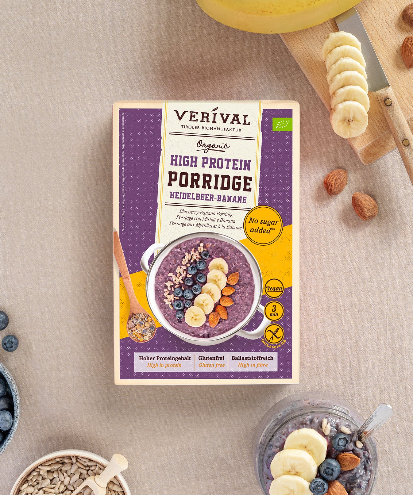 Ein Paket High Protein Porridge Heidelbeere-Banane von Verival Bio, umgeben von Bananenscheiben, Mandeln, Heidelbeeren und Haferflocken. 