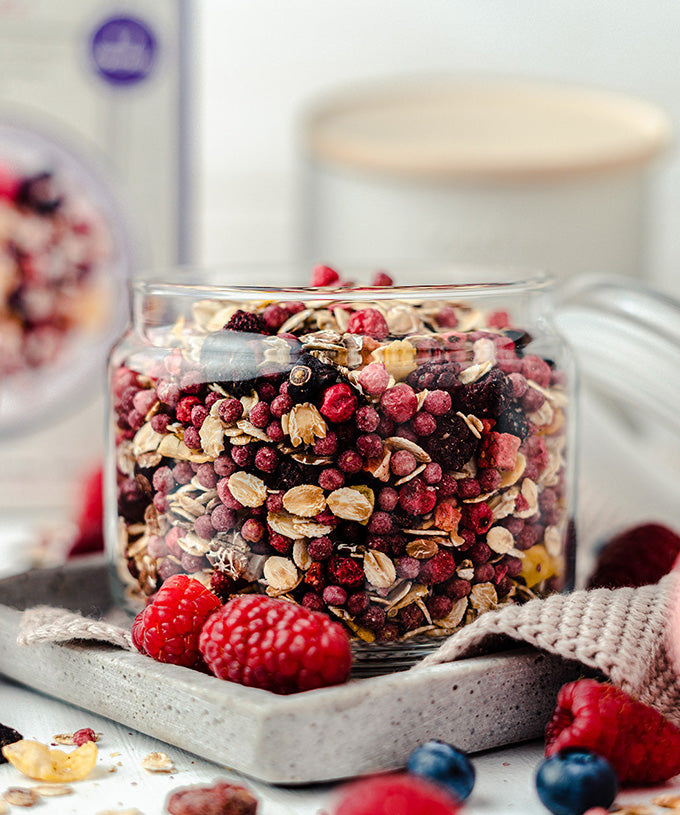 Auf einem Tablett steht ein Glas mit Bio Beeren Vollkorn Müsli von Verival Bio GmbH mit einer Mischung aus getrockneten Superfrüchten, umgeben von frischen Himbeeren und Heidelbeeren, während der Behälter und der Hintergrund unscharf sind.