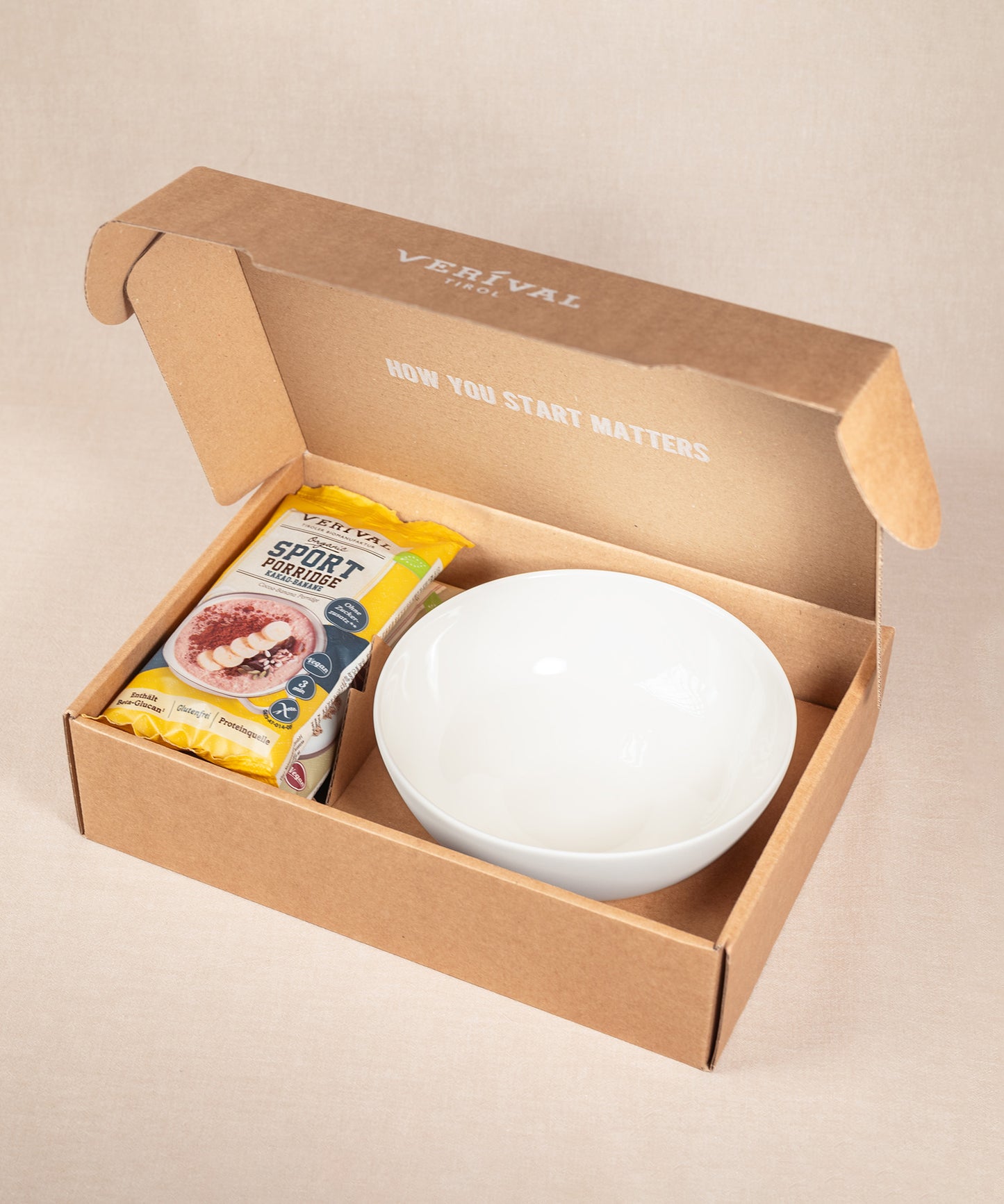 Die Geschenkverpackung für das Set „Breakfast Lover“ der Verival Bio GmbH besteht aus einer weißen Keramikschale und Sport Porridge im Hafer- und Fruchtdesign – ein ideales Geschenk für jeden Frühstücksliebhaber.
